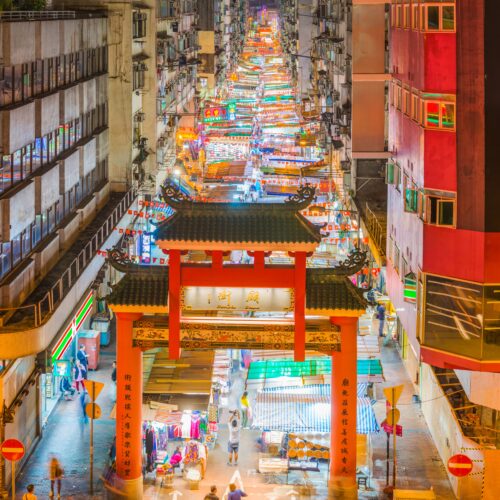 The highrise apartment buildings of Kowloon overlooking the crowded stalls of Temple Street Night Market and traditional Chinese entrance gate in Hong Kong.